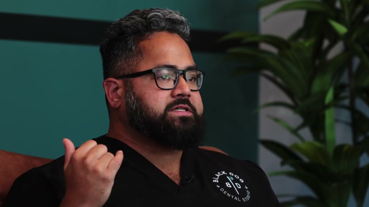 Man with glasses and a beard speaking, wearing a Black Dog Dental Studio uniform in an indoor setting with plants behind him.