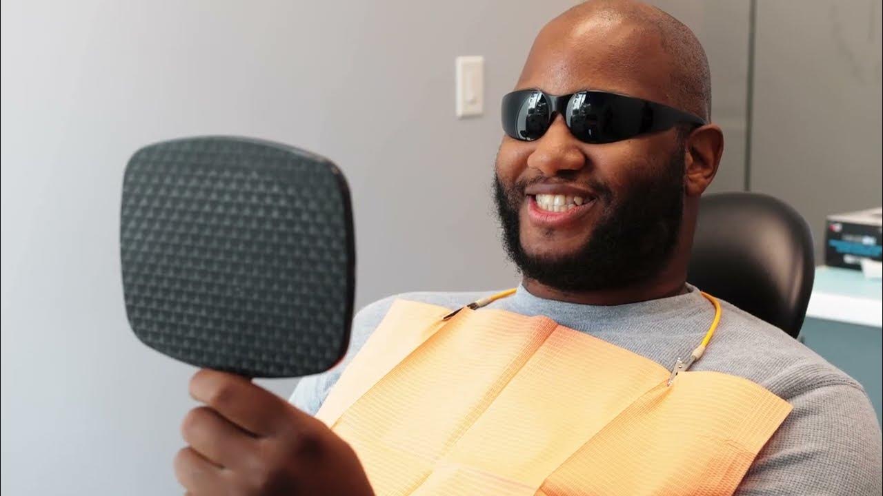 Smiling man wearing dark protective glasses and dental bib holding a hand mirror in a dental office.