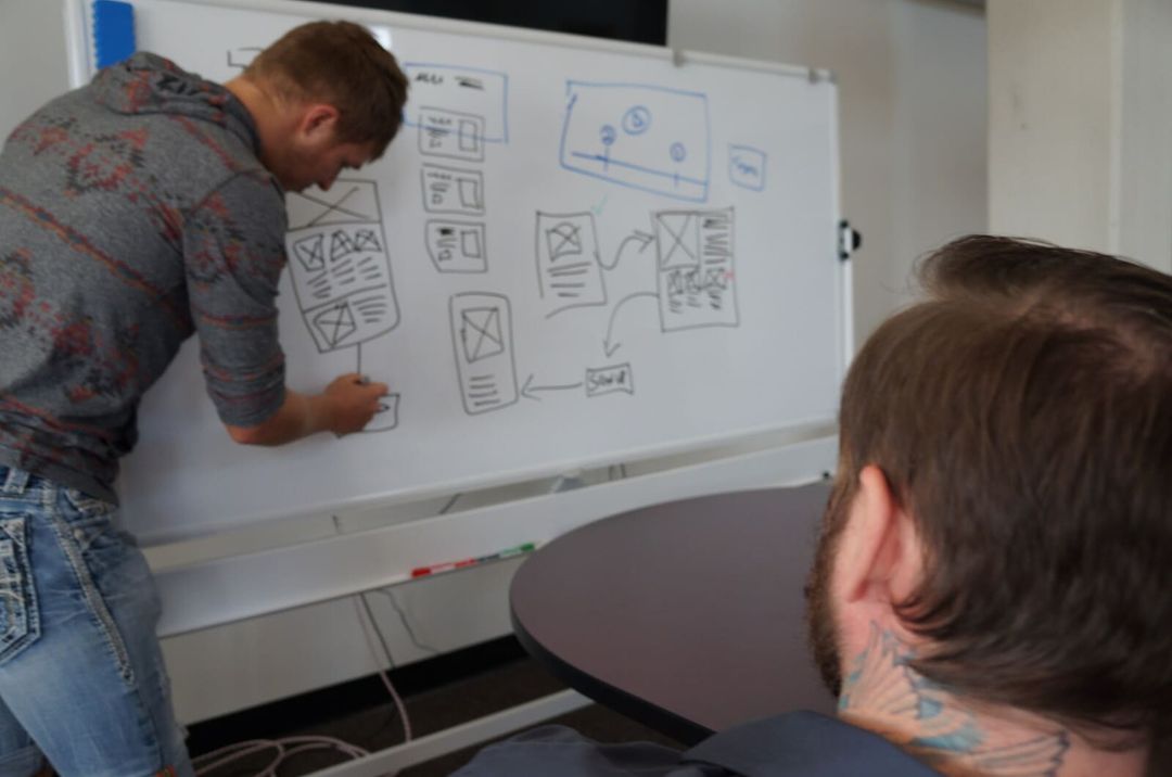 A VAULT engineer sketching app wireframes on a whiteboard while a colleague follows along