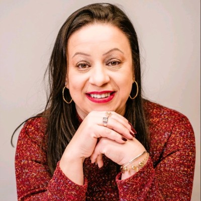 Mulher sorridente com cabelo escuro, brincos de argola e suéter vermelho, apoiando o queixo nas mãos.