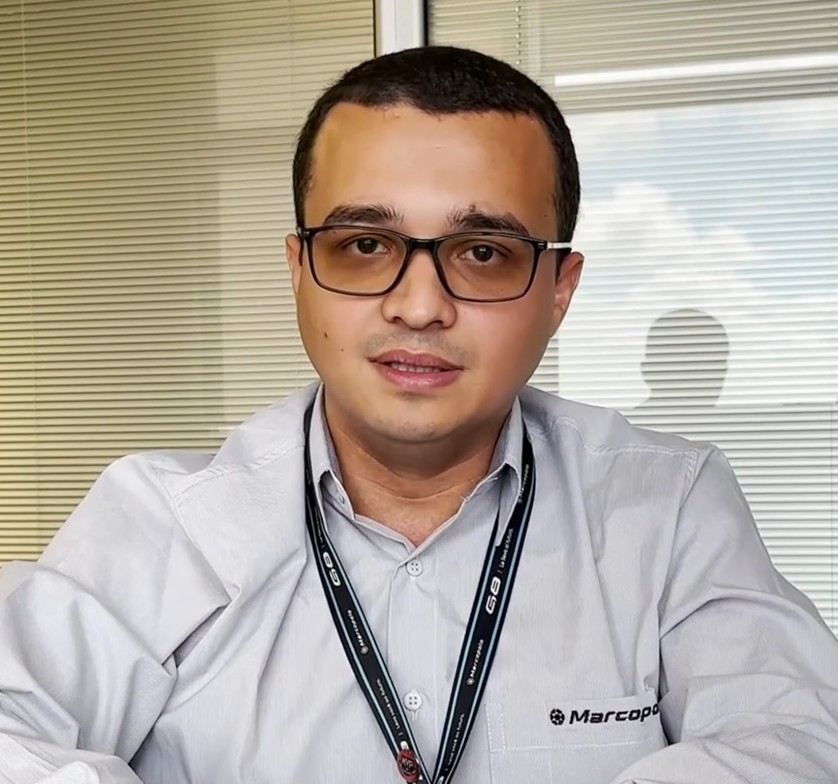 Homem de óculos e camisa cinza com crachá posando em frente a persianas e janela.