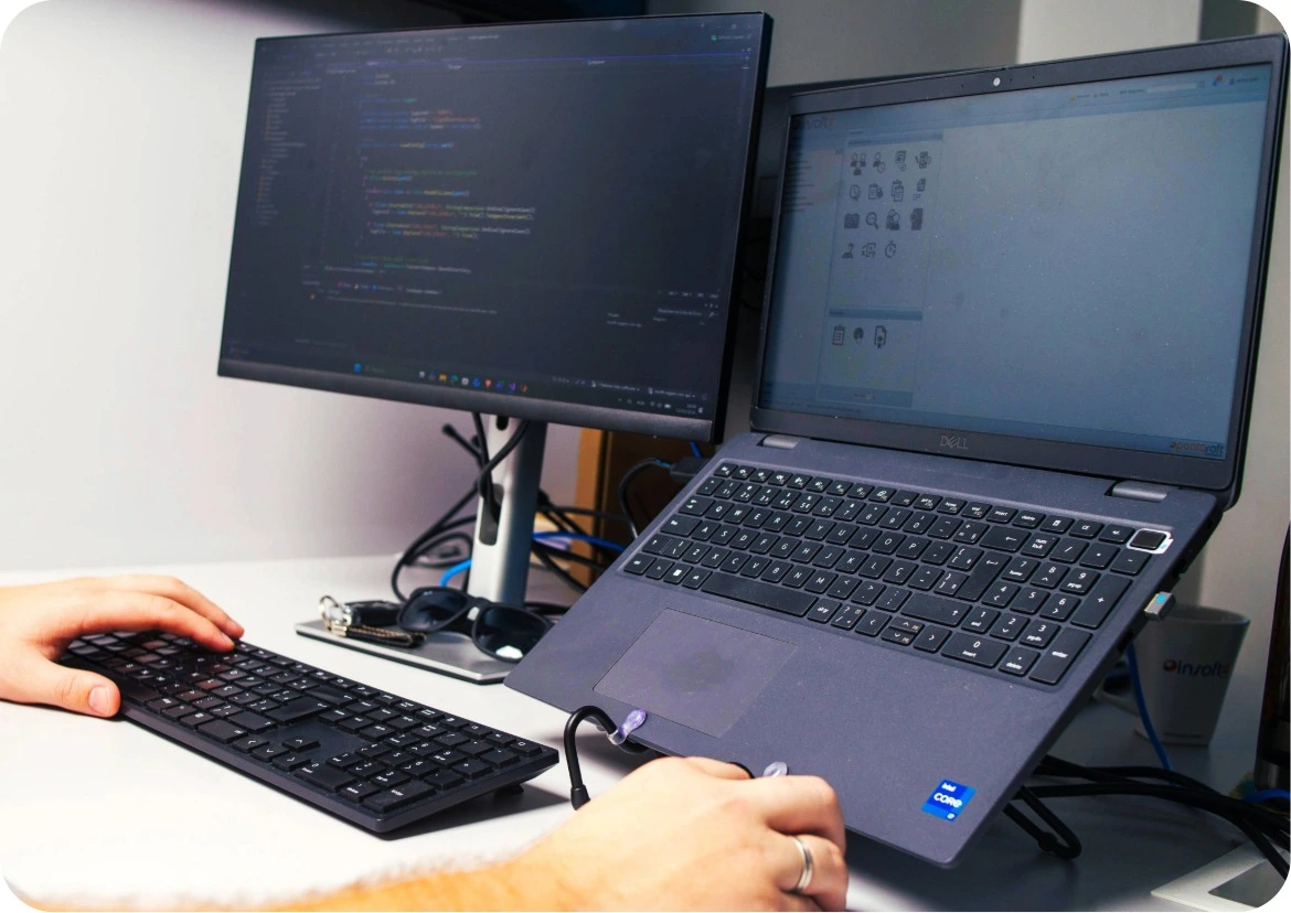 Pessoa usando teclado e mouse com laptop e monitor externo mostrando códigos na mesa de trabalho.