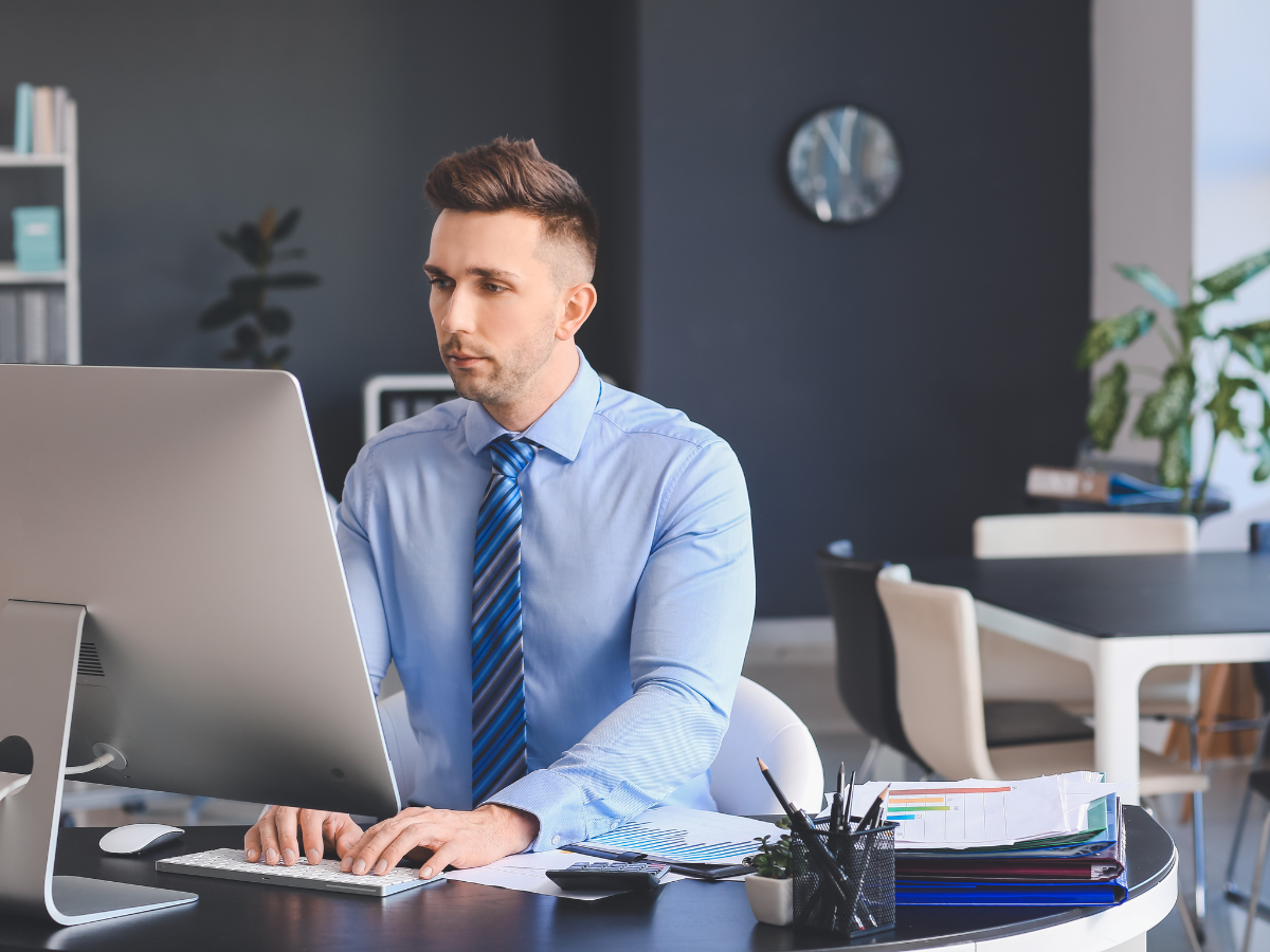 Man with gray hair and glasses working at desk with computer and papers in bright office.