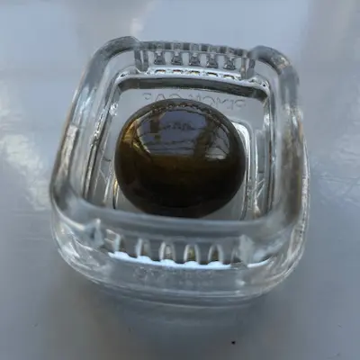 Glossy dark round old school cannabis hash ball placed inside a clear square glass container on a reflective surface.