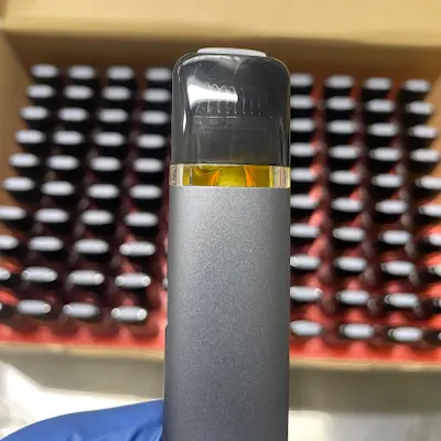 Close-up of a vape cartridge filled with light amber hash rosin attached to a gray vape pen, with a blurred background of multiple similar cartridges in a red tray.