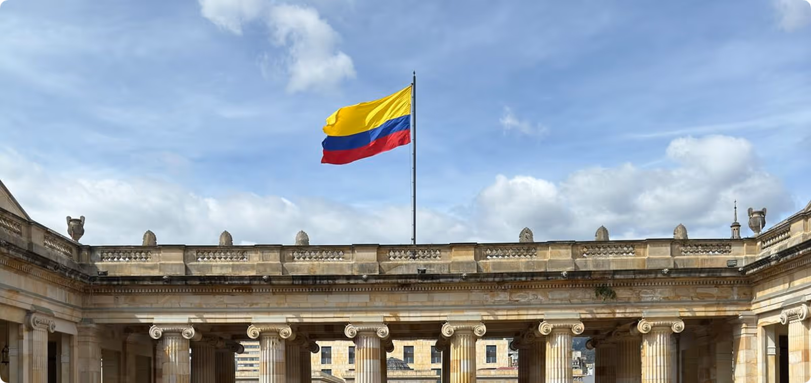 The national flag of Colombia flying prominently above the neoclassical stone columns of the Palace of Justice at Bolívar Square in Bogotá under a bright, cloudy sky.