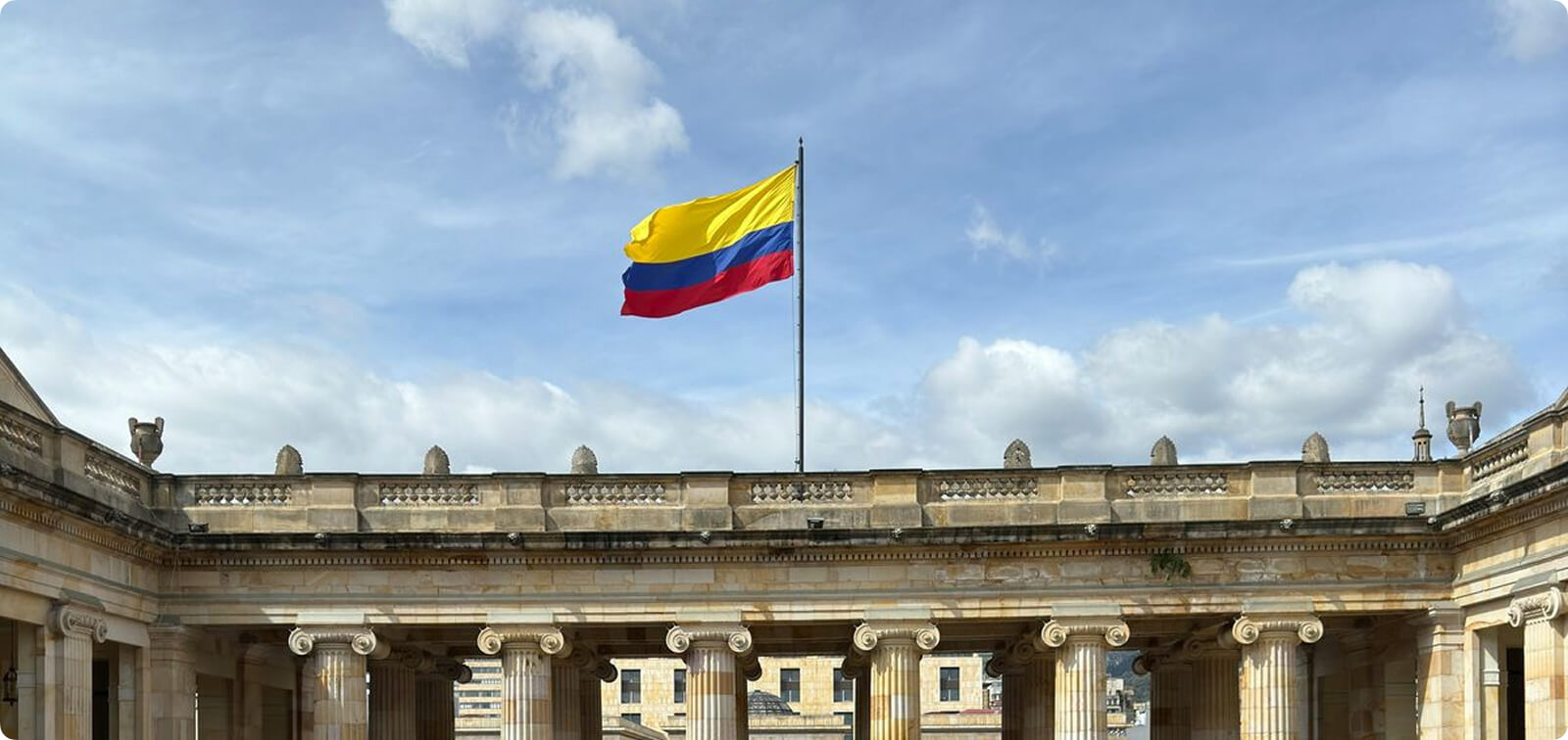 The national flag of Colombia flying prominently above the neoclassical stone columns of the Palace of Justice at Bolívar Square in Bogotá under a bright, cloudy sky.