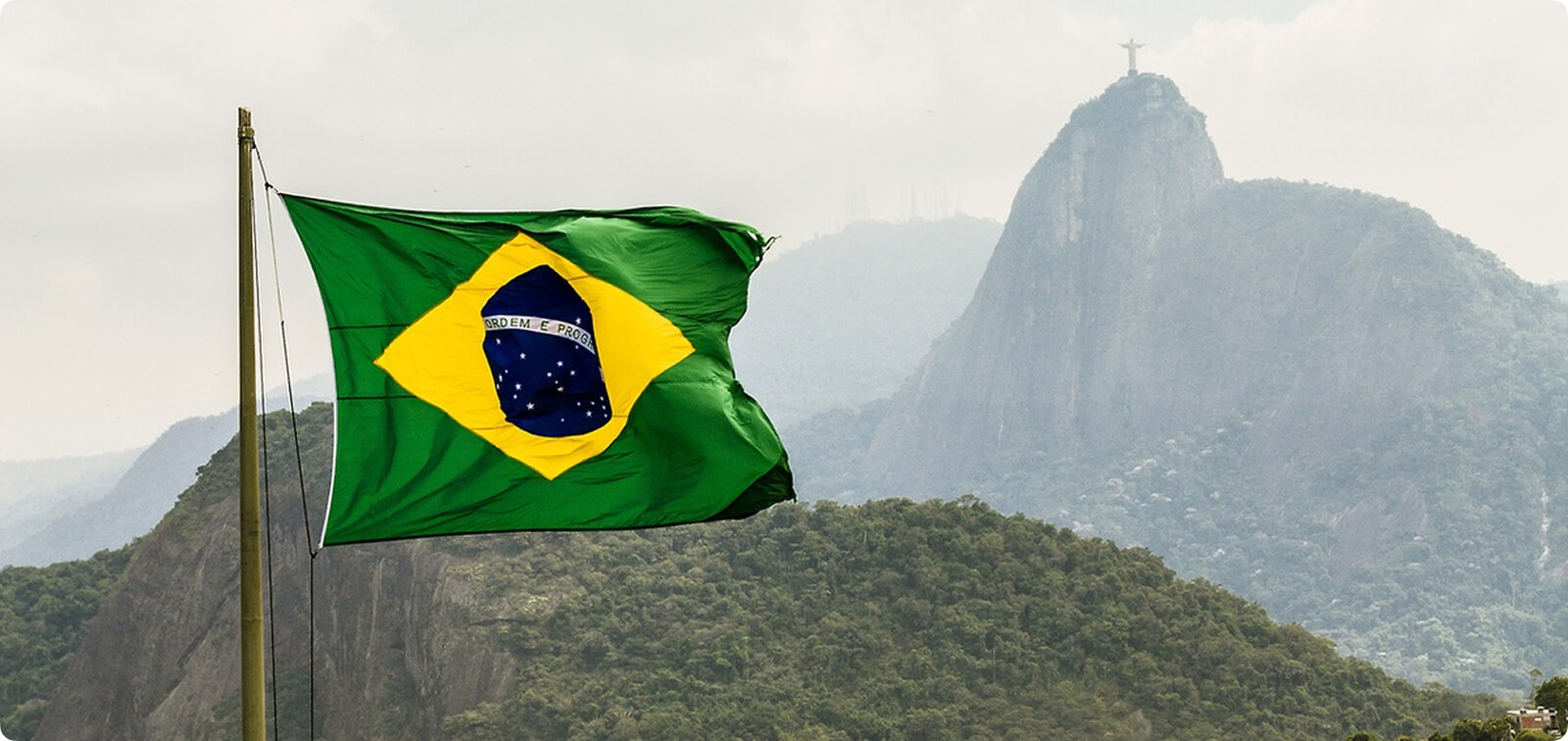 The Brazilian flag waving in the wind with the iconic Christ the Redeemer statue on Corcovado Mountain and the Rio de Janeiro landscape in the background.