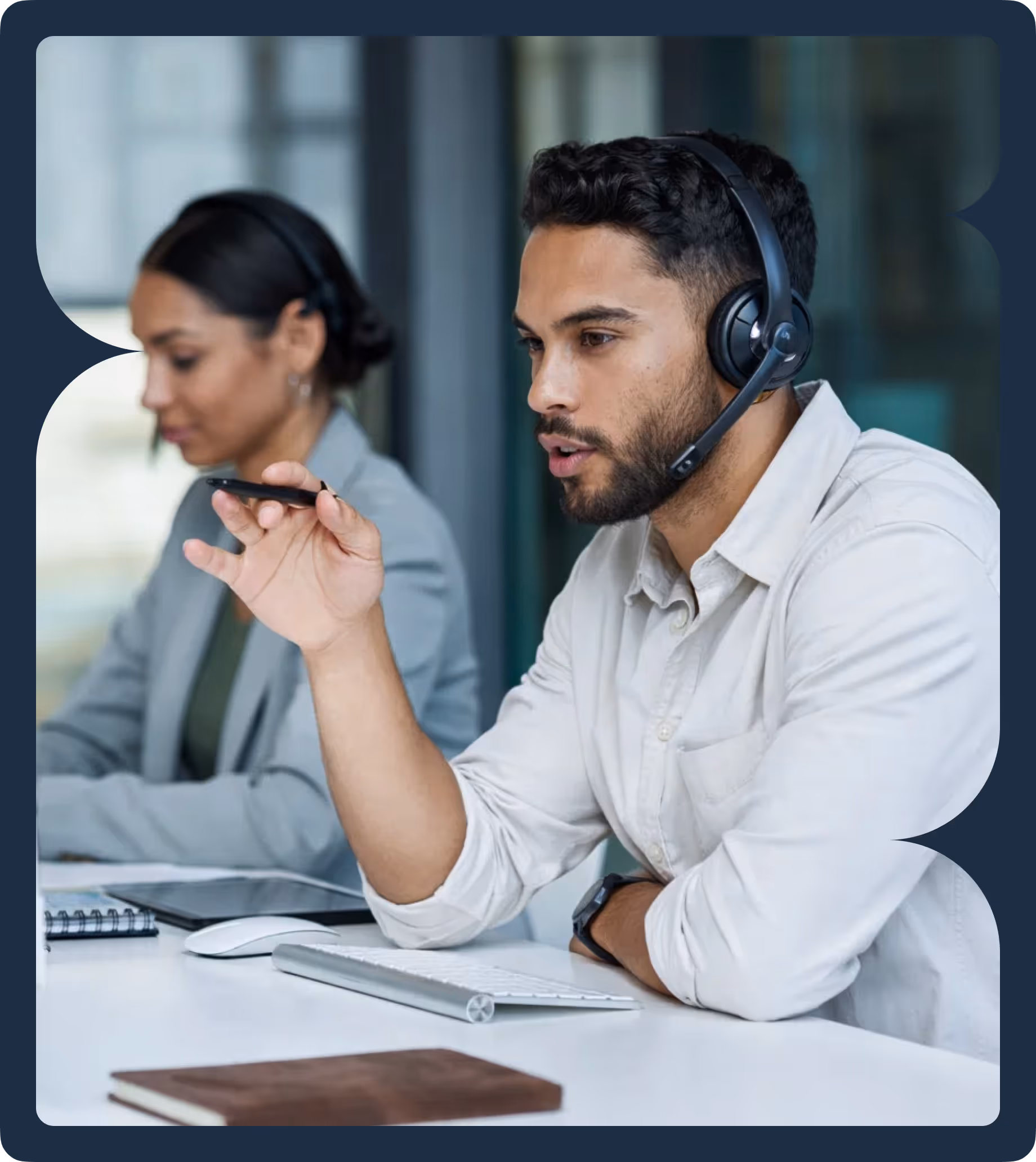 Man wearing a headset speaking and gesturing with a pen while a woman wearing a headset works in the background.