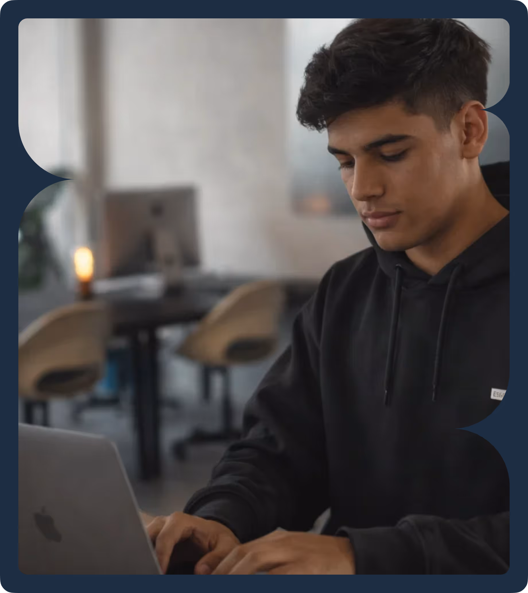 Young man in a black hoodie focused on typing on a laptop in a modern office space.