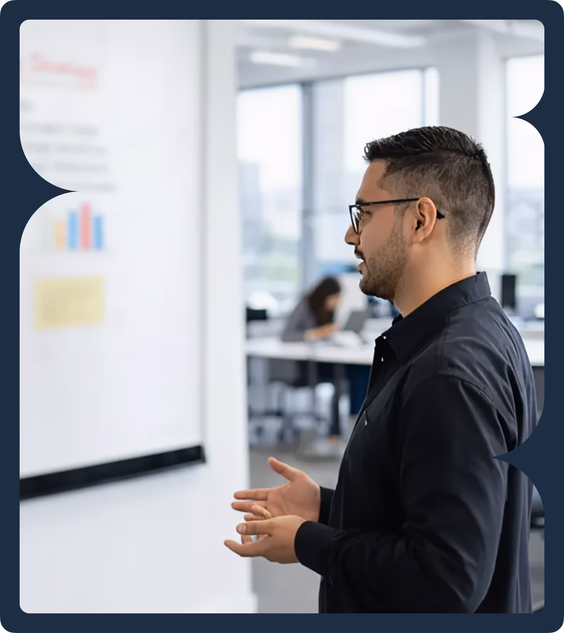 Man with glasses and a beard giving a presentation in an office, gesturing with hands toward a whiteboard with charts.
