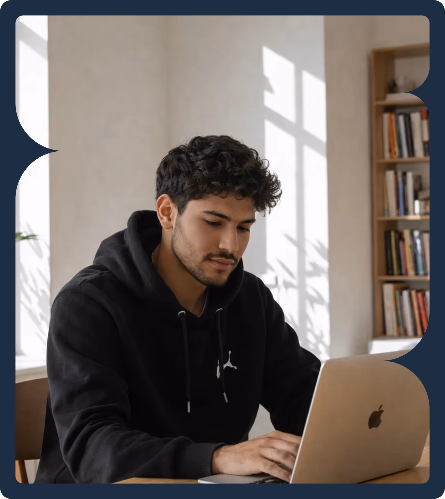 Man in black hoodie working on a silver Apple laptop at a wooden desk in a room with bookshelf and sunlight streaming through the window.