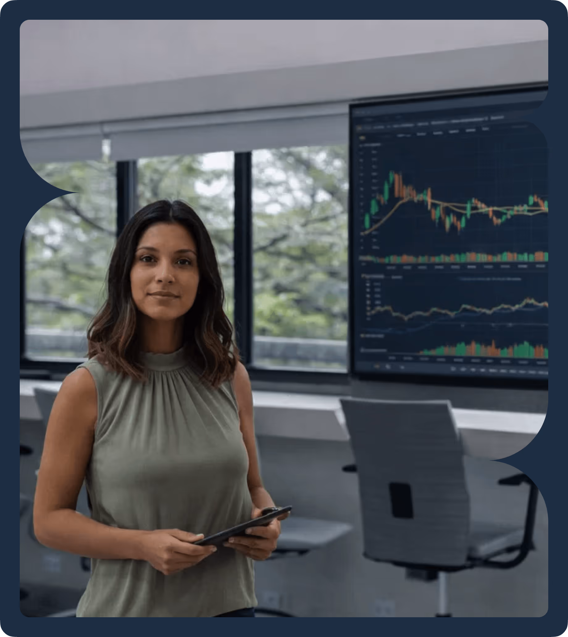 Woman holding a tablet standing in a modern office with a large screen displaying stock market charts in the background.