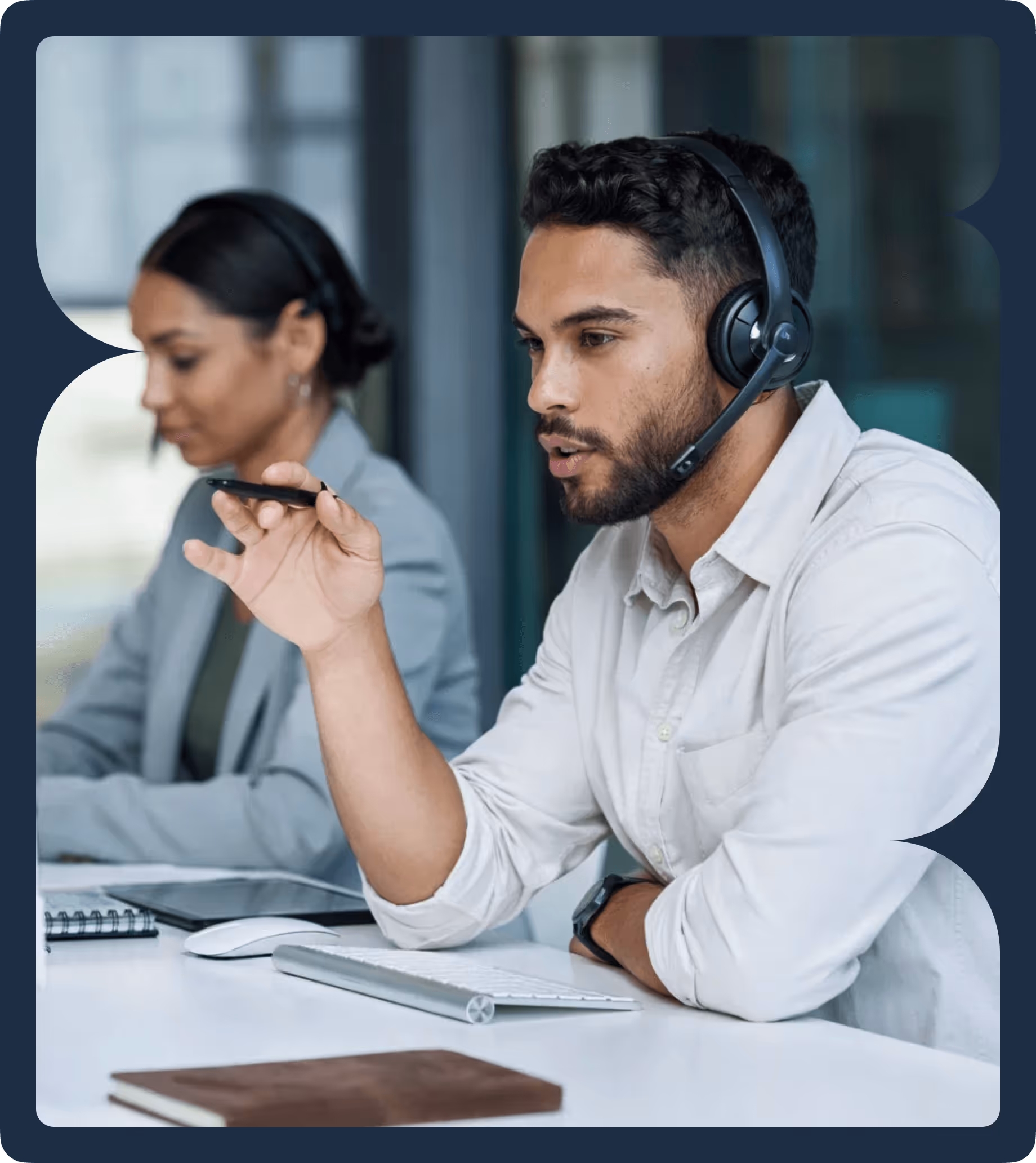 Man wearing headset and holding a pen, speaking while working at a desk with keyboard, mouse, and notebook, with a woman wearing a headset working in the background.
