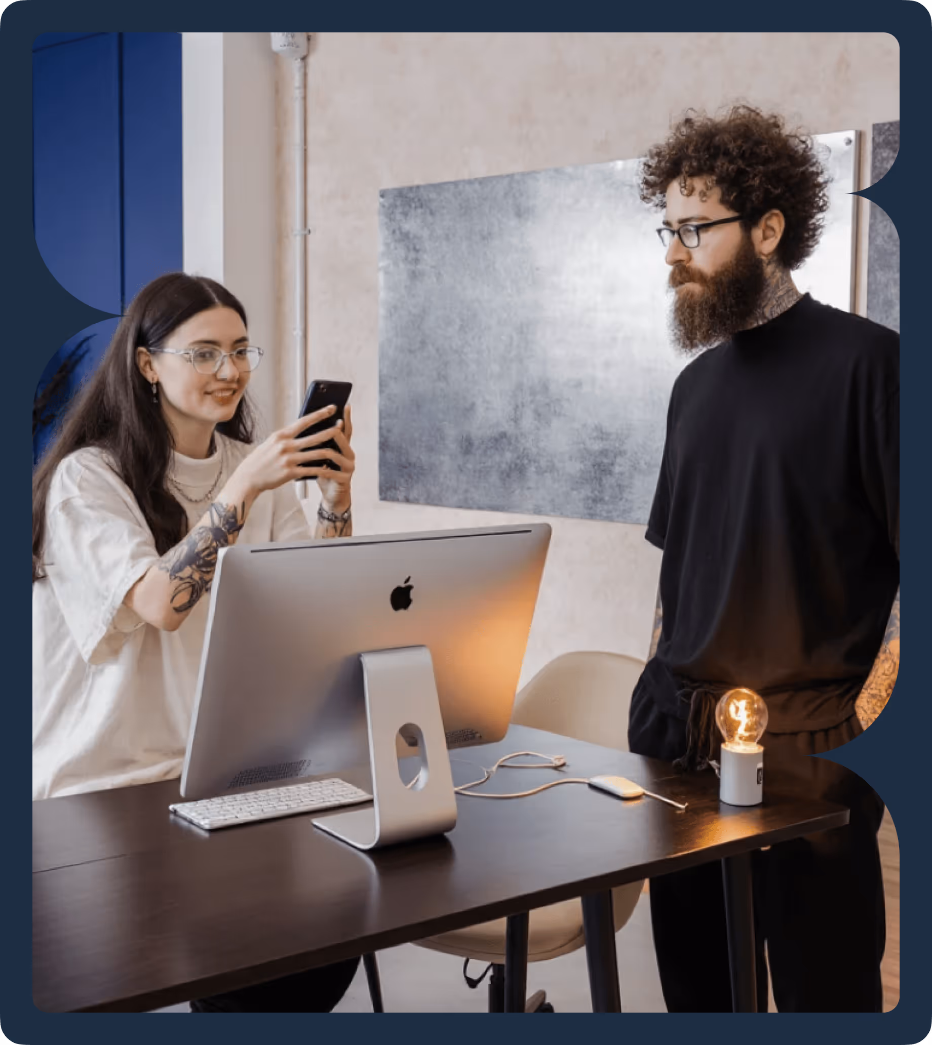 Two people with tattoos working together at a desk with an Apple computer, one taking a photo with a smartphone, the other standing beside a lit light bulb.