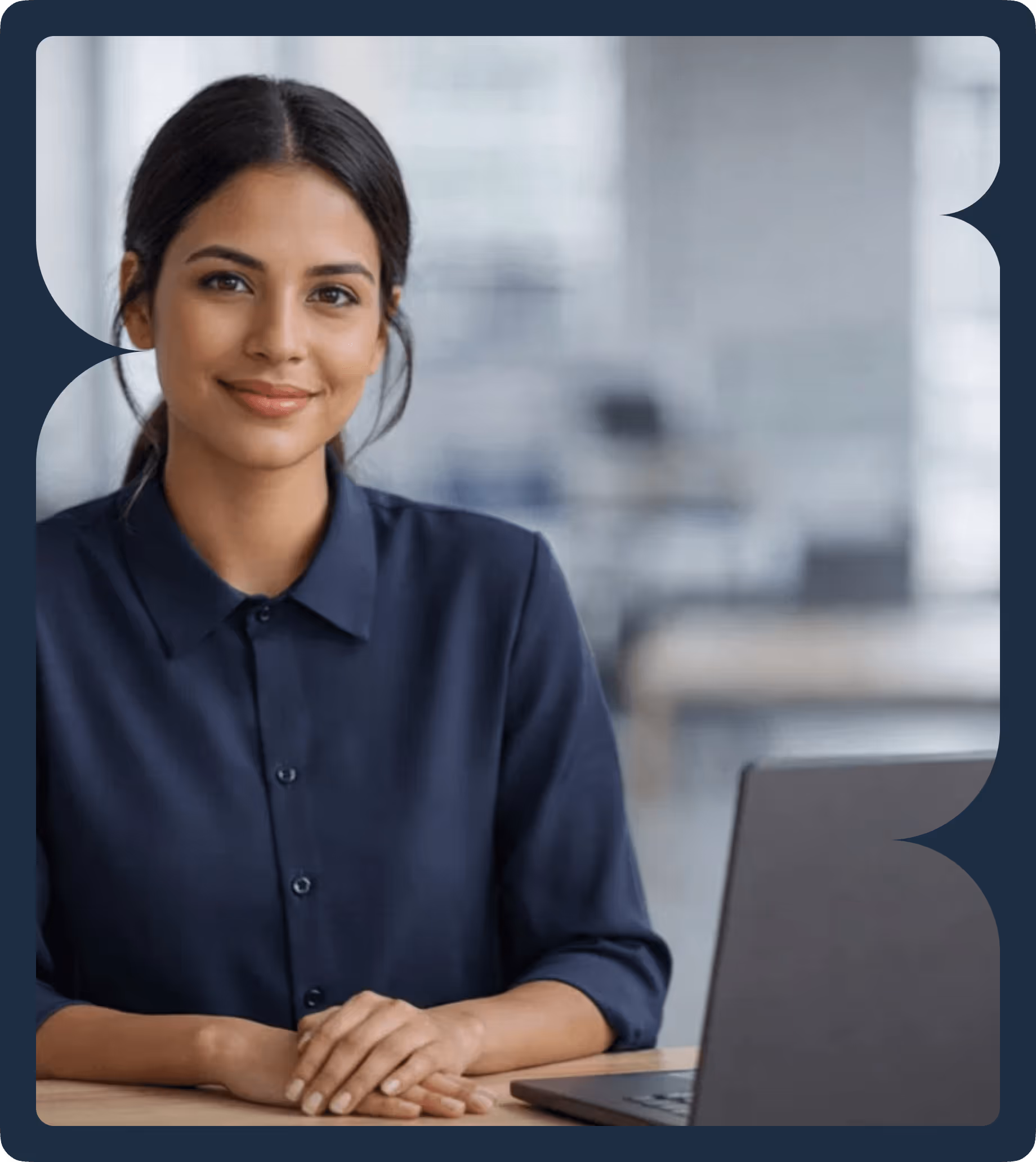 Smiling woman with dark hair wearing a navy blue shirt, sitting at a desk with a laptop in an office setting.