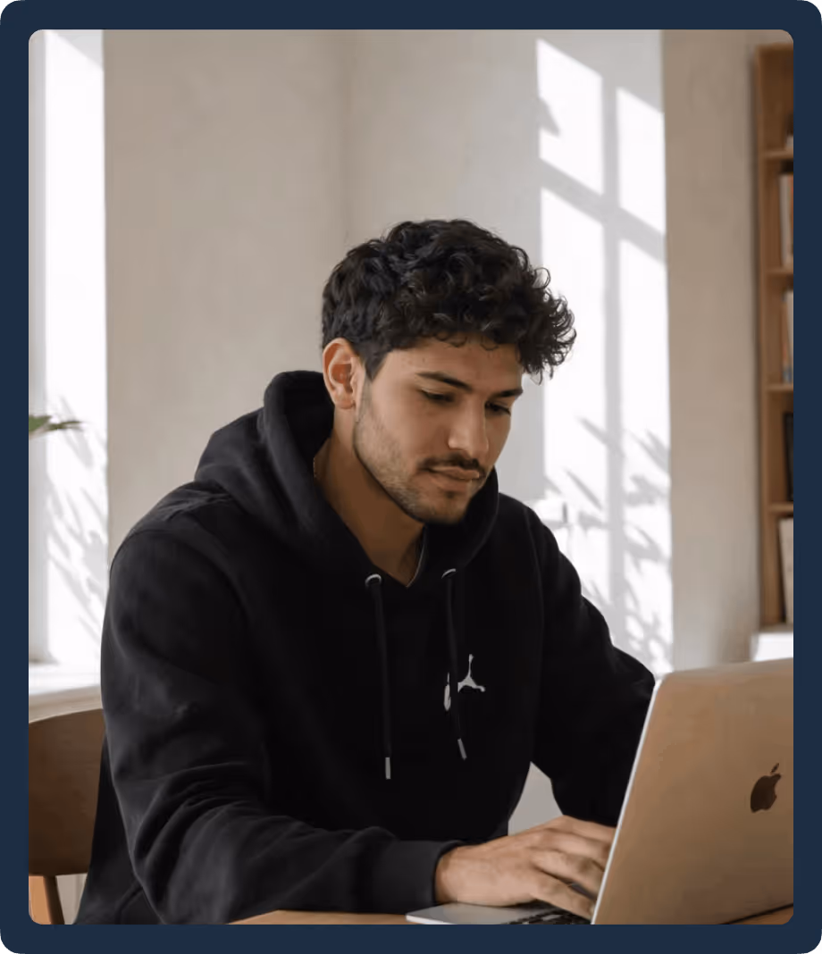 Young man with curly hair wearing a black hoodie working on a silver MacBook laptop in a bright room.