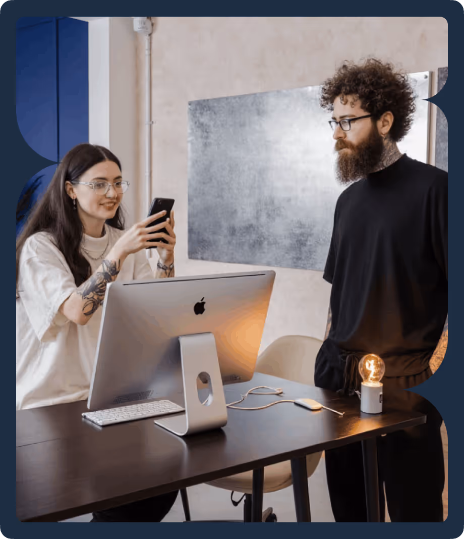 Two people with glasses at a desk; one woman is holding a smartphone, and a man with a beard stands beside a computer and a lit bulb on the desk.