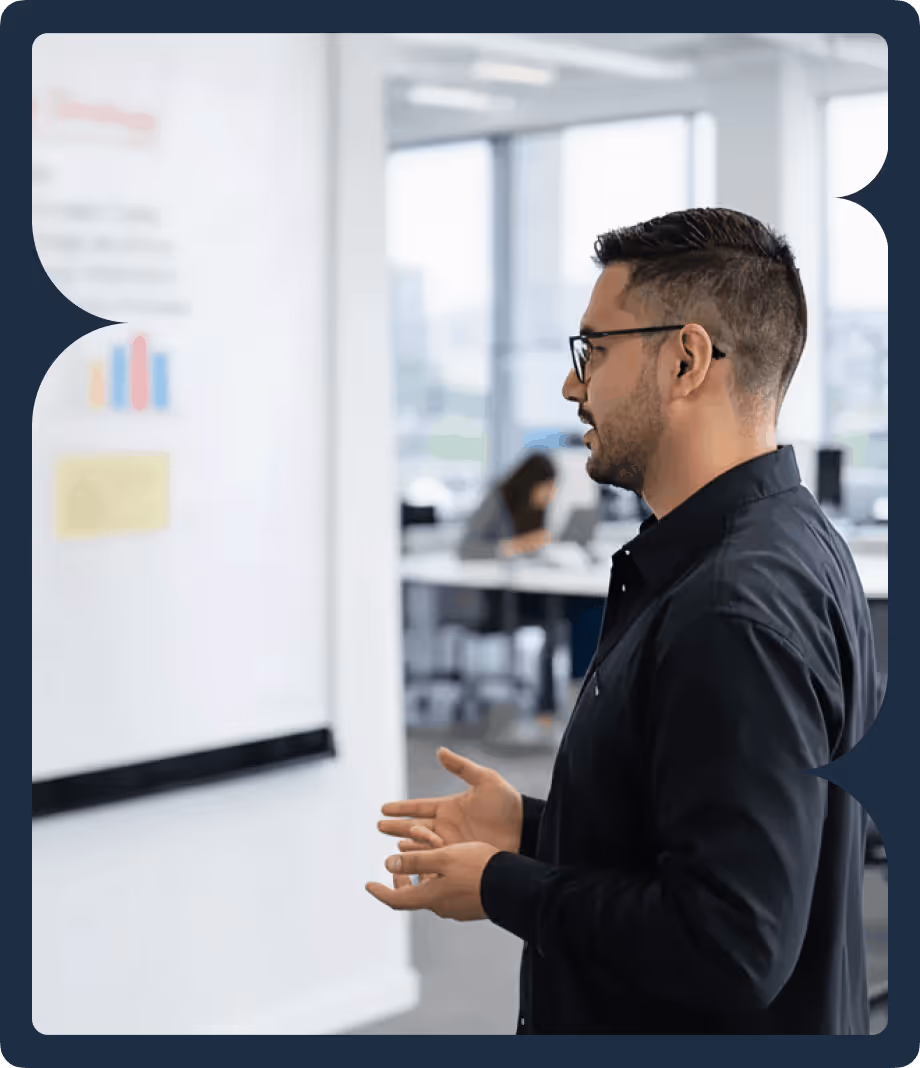 Man wearing glasses and a black shirt giving a presentation in an office, gesturing with his hands near a whiteboard with charts.
