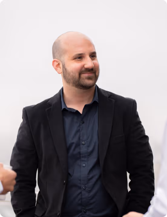 Smiling bald man with beard wearing a black blazer and dark shirt standing in a light background.
