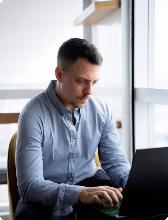 Man in blue shirt typing on a laptop by a large window with city view.