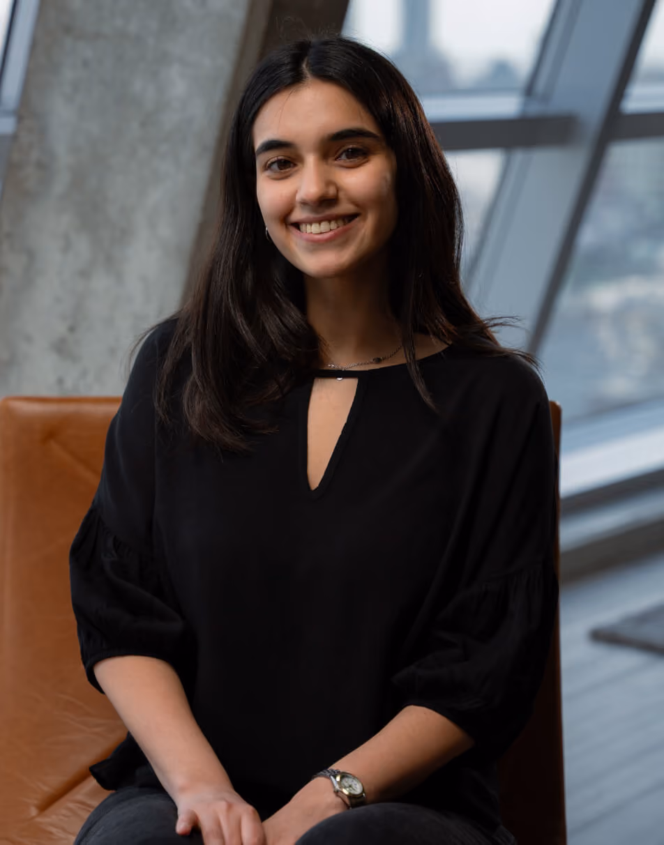 Smiling young woman with long dark hair wearing a black top, sitting on a brown chair.