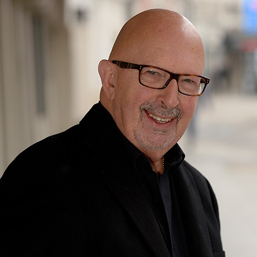 Smiling bald man with glasses wearing a black coat and black shirt, standing outdoors with a blurred background.