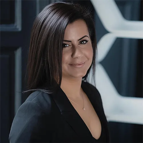 Woman with straight dark brown hair wearing a black blazer and smiling slightly, with geometric patterns in the blurred background.