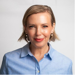 Woman with short light brown hair wearing pearl earrings and a light blue buttoned shirt, smiling against a plain light background.