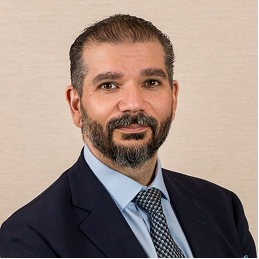 Man with short dark hair and beard wearing a dark suit jacket, light blue shirt, and patterned tie, posing against a plain beige background.