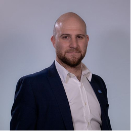 A professional man with a shaved head and beard wearing a navy blue blazer over a white shirt, posing against a plain light grey background.