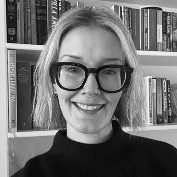 Smiling woman with short blonde hair and large black glasses wearing a black top, standing in front of a bookshelf filled with books.