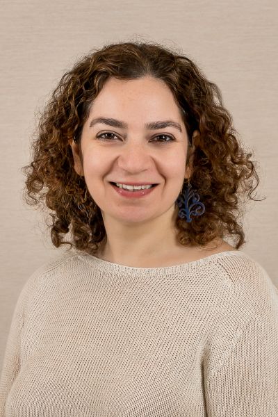 Woman with curly brown hair smiling, wearing a cream knitted sweater and blue fleur-de-lis earrings against a beige background.