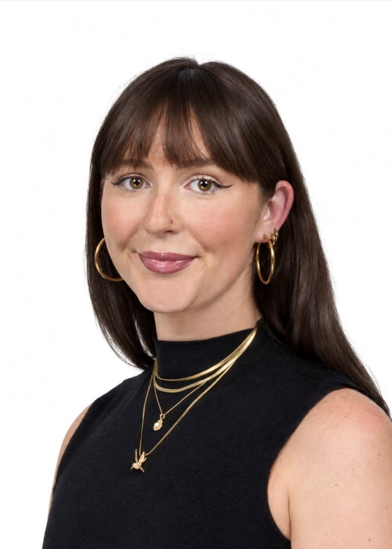 Woman with brown hair and bangs wearing a black sleeveless top, gold hoop earrings, and layered gold necklaces, smiling against a white background.