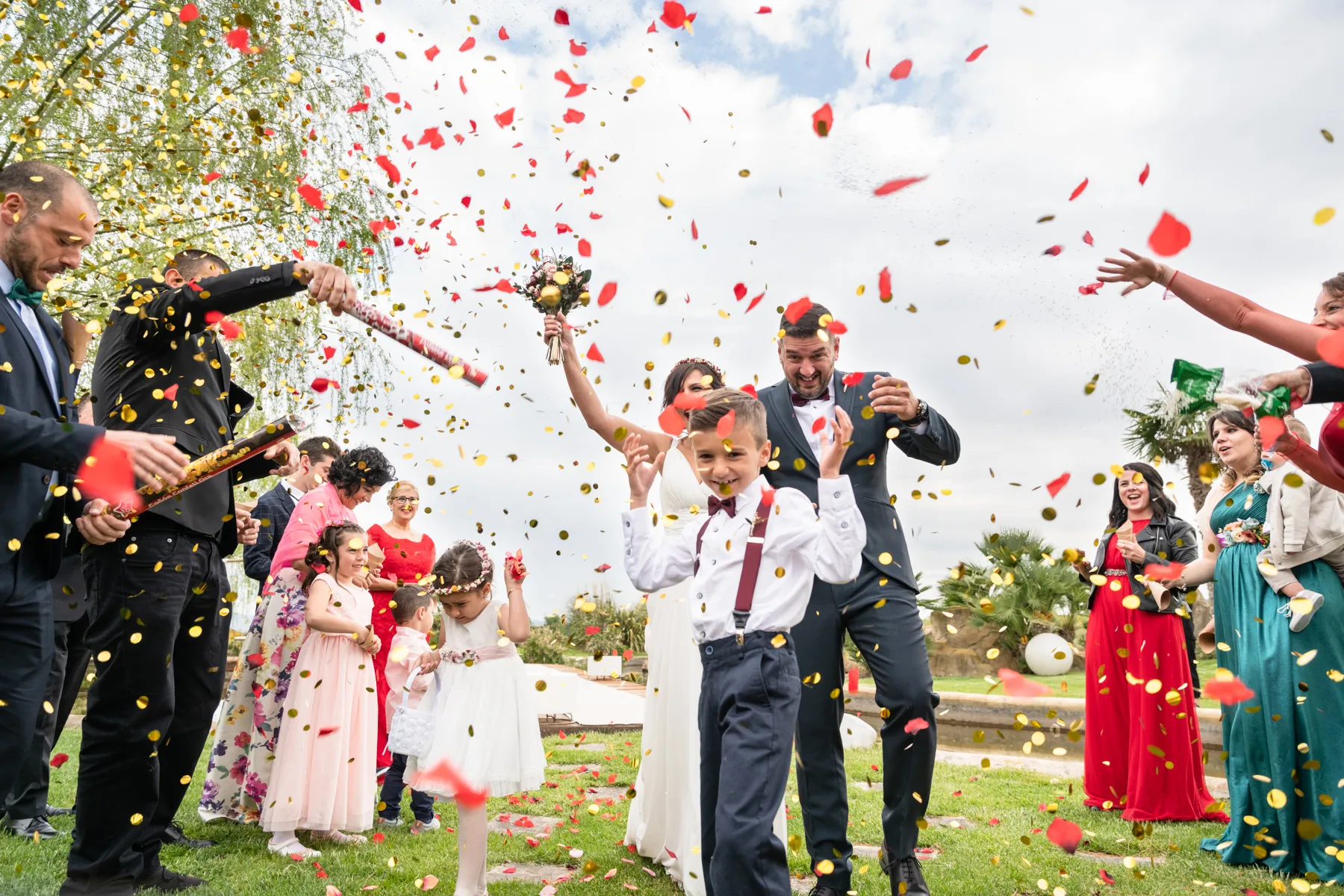 Novia, novio, niños e invitados celebrando al aire libre con confeti rojo y dorado en una boda.