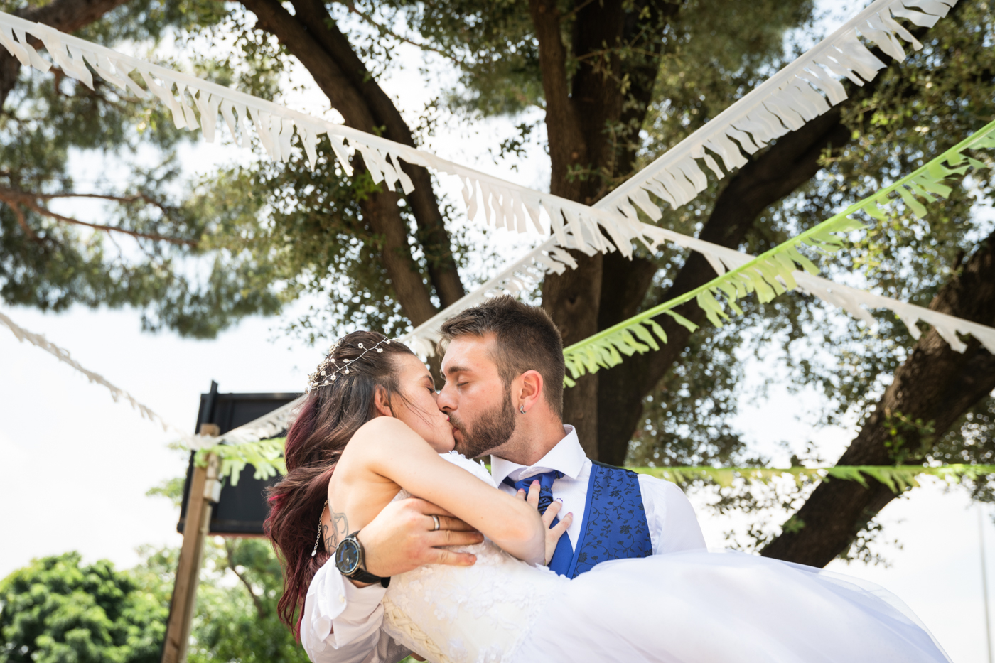 Novio con chaleco azul sosteniendo y besando a la novia con vestido blanco al aire libre, bajo serpentinas de papel.