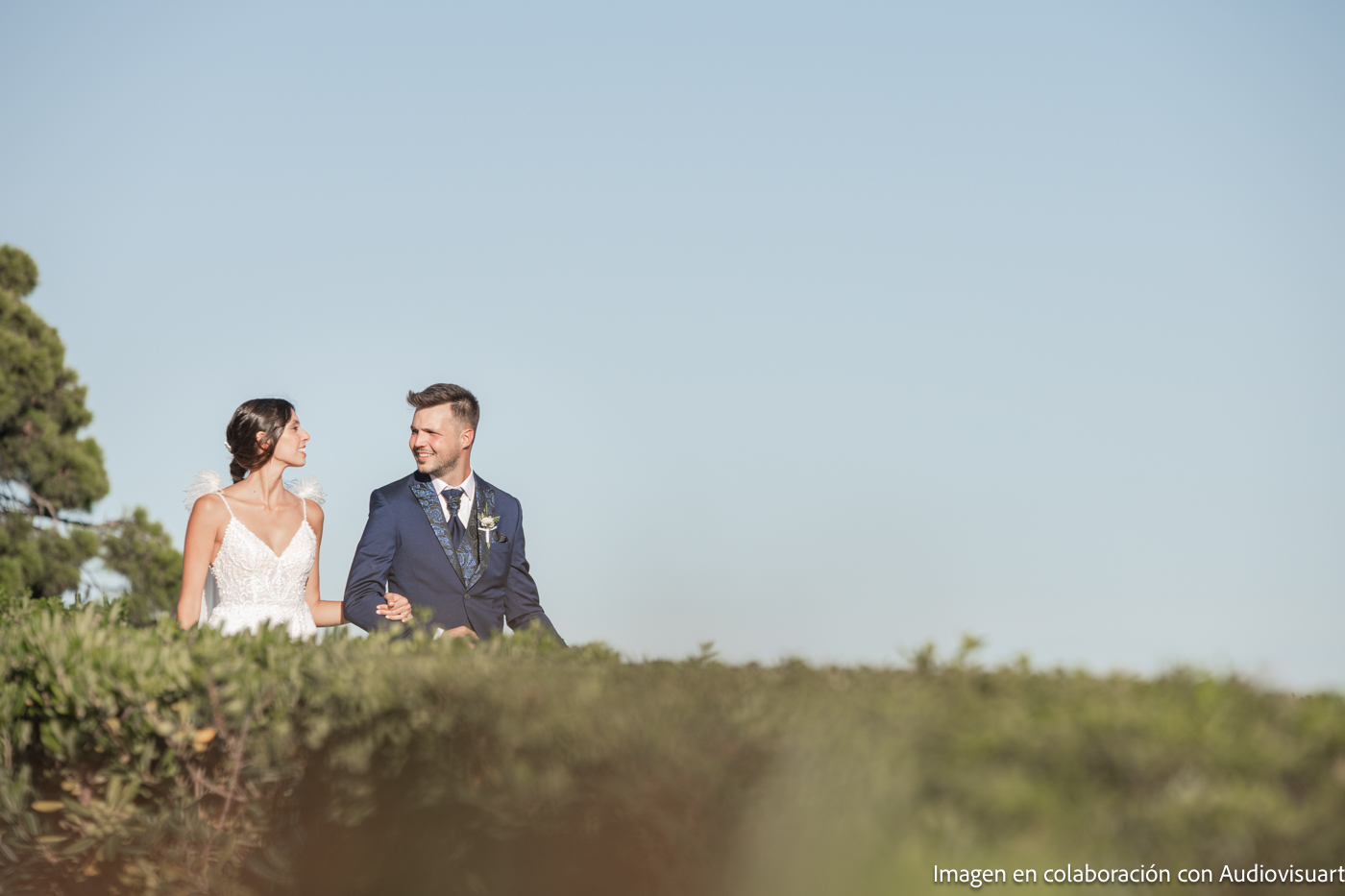 Novia y novio tomados de la mano y mirándose al aire libre, con cielo azul despejado y vegetación en primer plano.