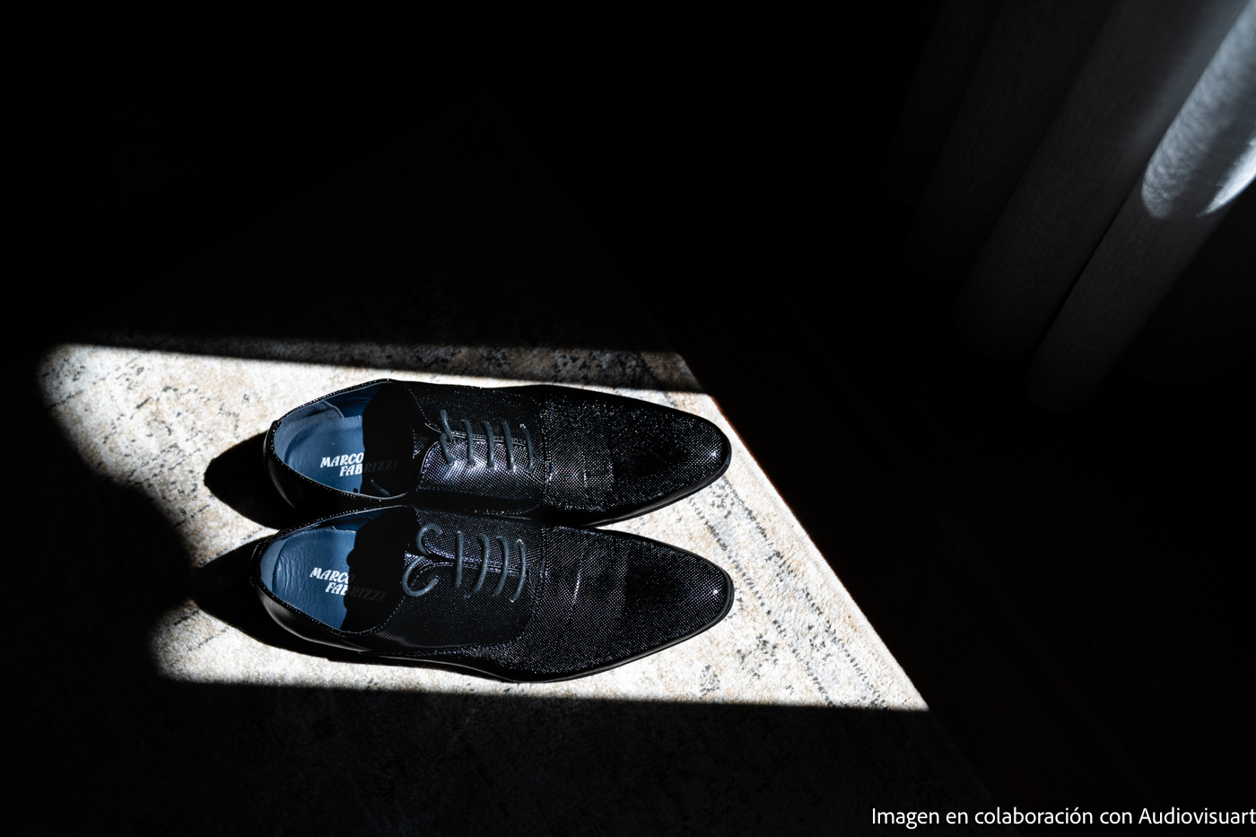 Par de zapatos de vestir negros con cordones colocados uno al lado del otro sobre una alfombra, iluminados por un haz de luz rectangular.