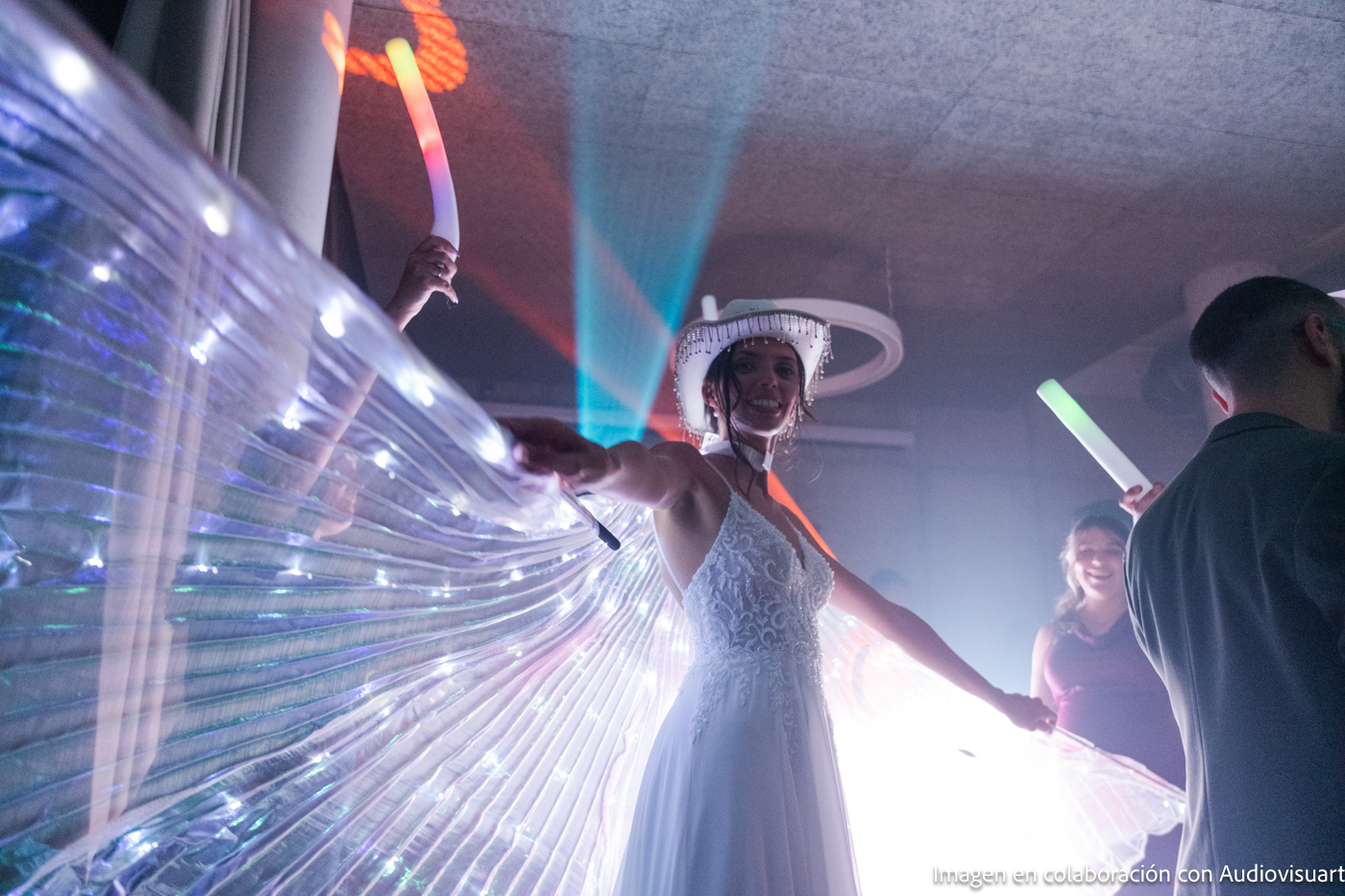 Novia con vestido blanco y alas de ángel iluminadas bailando con invitados que sostienen varas luminosas en una fiesta.