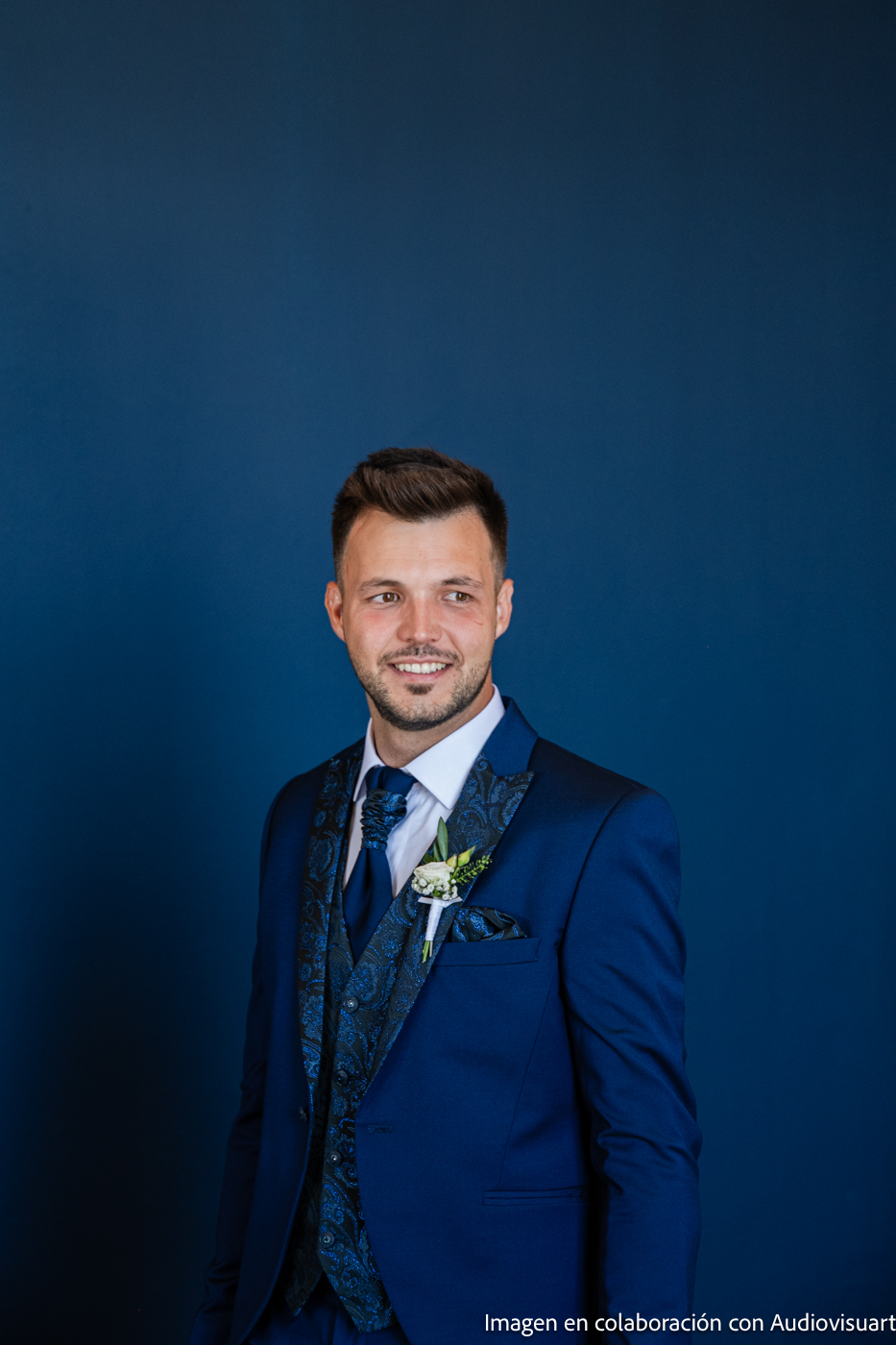 Hombre sonriente con traje azul, chaleco estampado y boutonnier contra un fondo azul sólido.