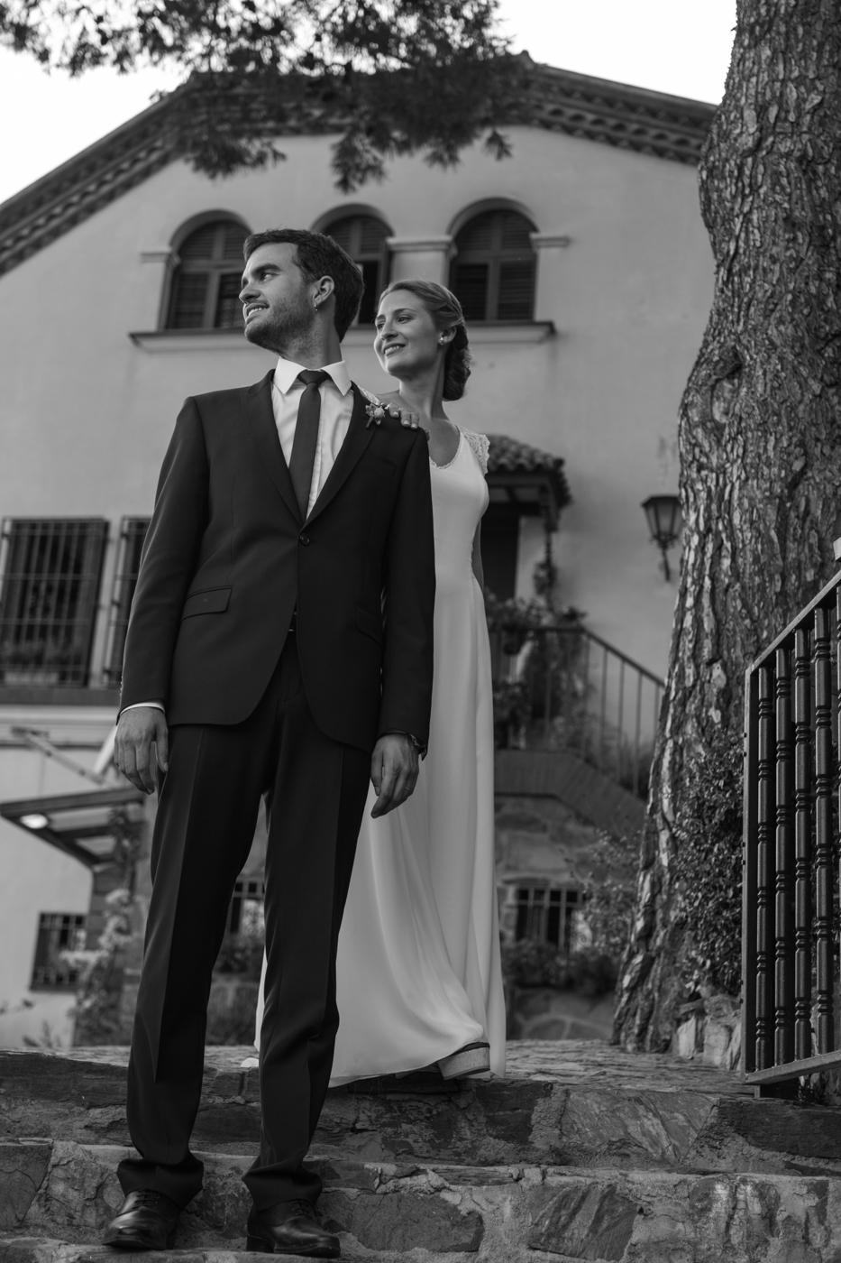 Novia con vestido blanco de pie detrás del novio con traje oscuro en unos escalones de piedra frente a un edificio rústico; ambos miran hacia un lado y sonríen.
