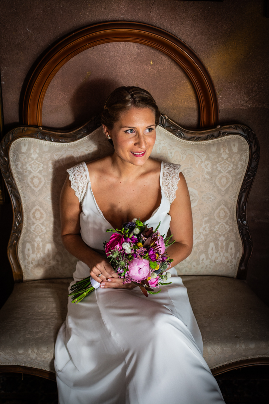 Novia sonriente con vestido blanco, sosteniendo un ramo de flores colorido, sentada en un sofá vintage ornamentado.