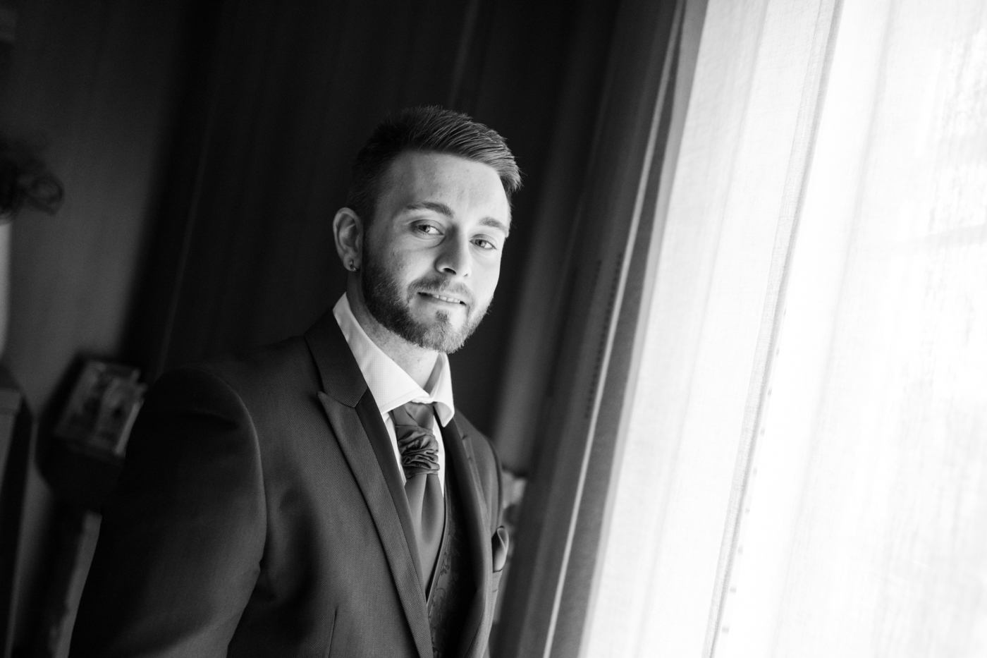 Hombre joven con barba, vestido con traje y corbata, de pie junto a una ventana luminosa y sonriendo suavemente.