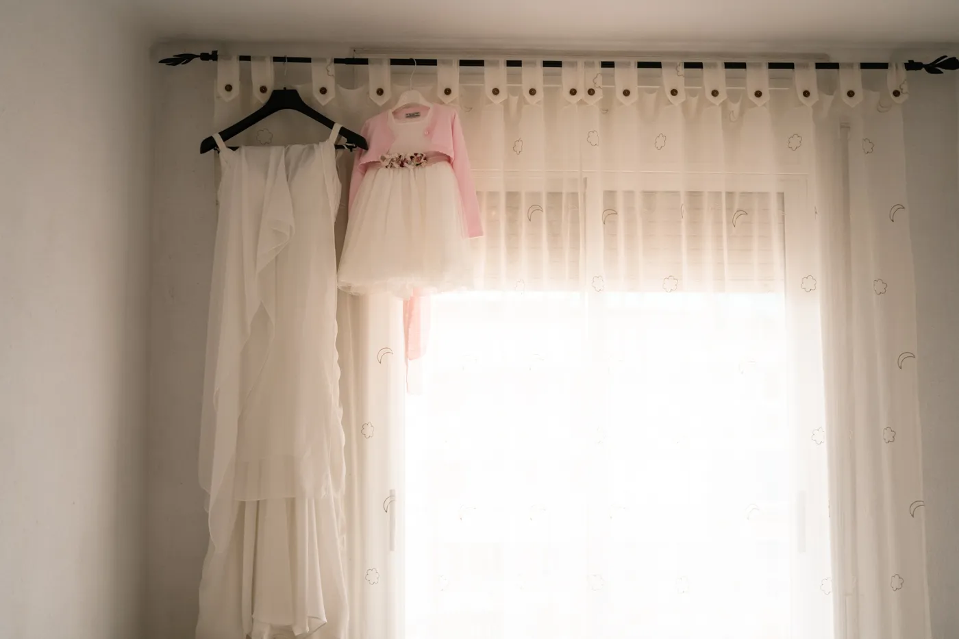 Vestido blanco de adulto y vestido blanco de niña con rebeca rosa colgados en perchas negras frente a una cortina translúcida con estampados.