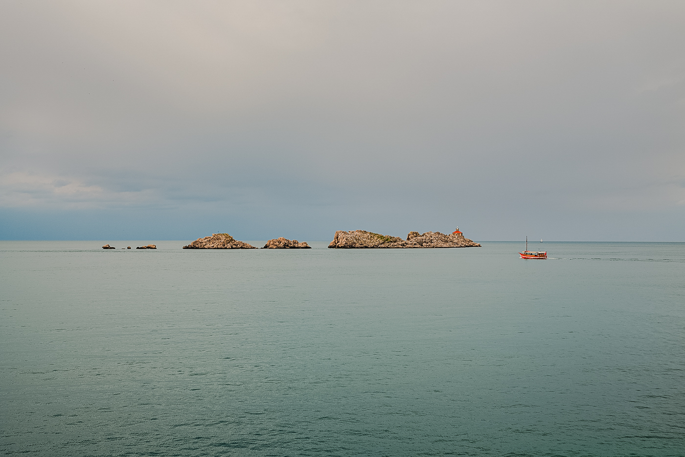 Mar en calma con pequeñas islas rocosas y una barca roja navegando cerca de la isla más grande bajo un cielo nublado.