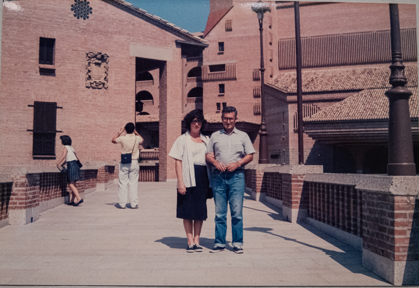 Una pareja posando del brazo en un amplio paseo de piedra, con edificios de ladrillo y farolas al fondo en un día soleado.
