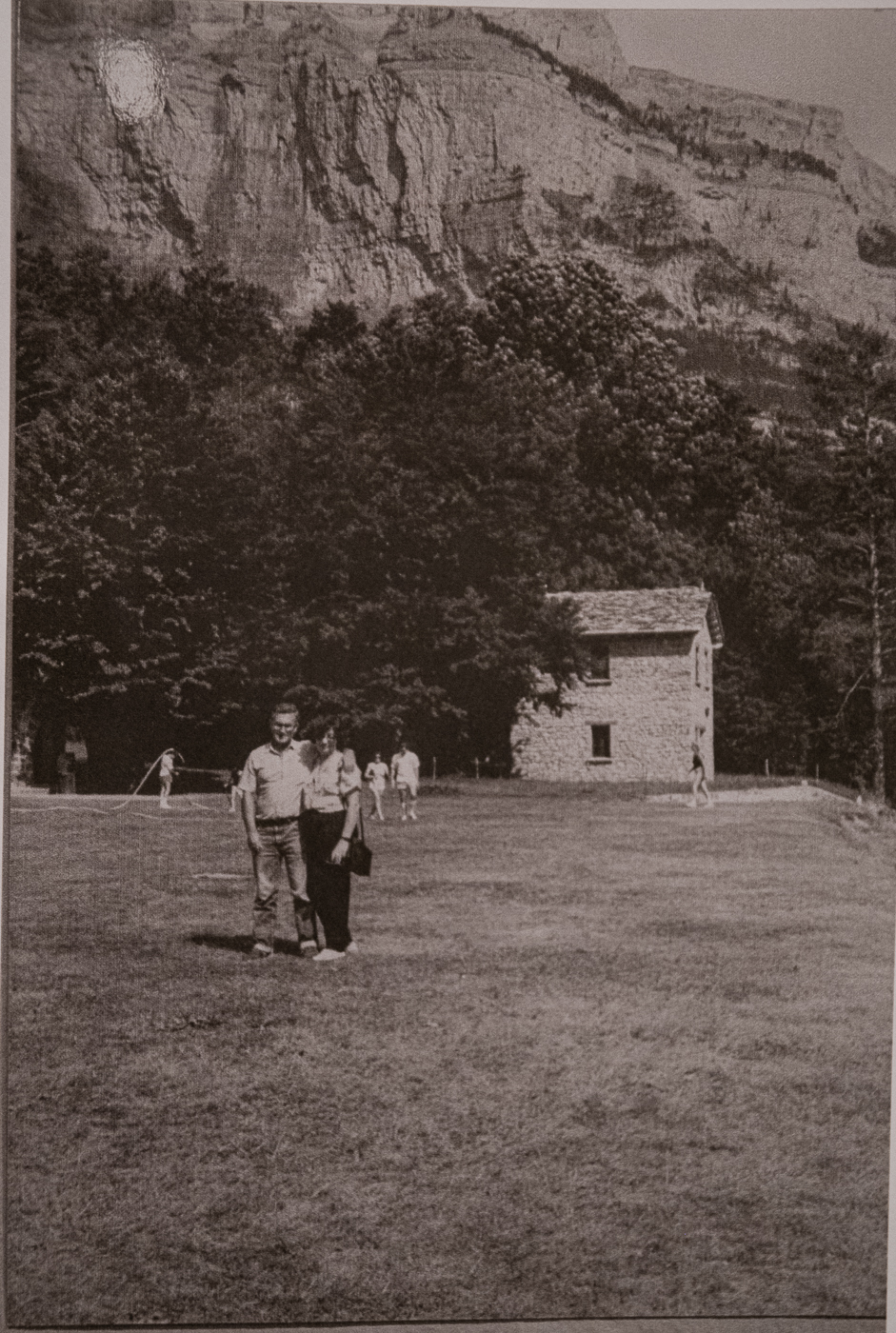 Foto en blanco y negro de una pareja de pie en un campo de hierba, con una pequeña casa de piedra y acantilados montañosos cubiertos de bosque al fondo.