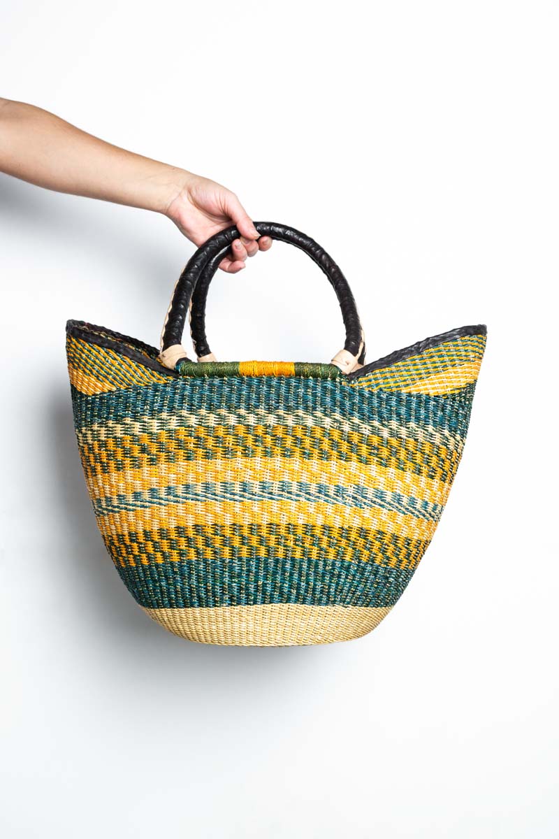 Mano sosteniendo un bolso de rafia con rayas amarillas, verdes y color natural y con asas negras. 