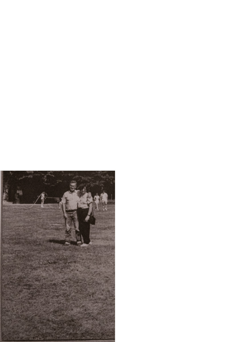 Foto vintage en blanco y negro de una pareja sonriente de pie al aire libre, con árboles y personas al fondo.