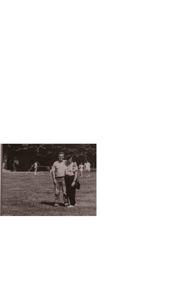 Foto vintage en blanco y negro de una pareja sonriente de pie al aire libre, con árboles y personas al fondo.